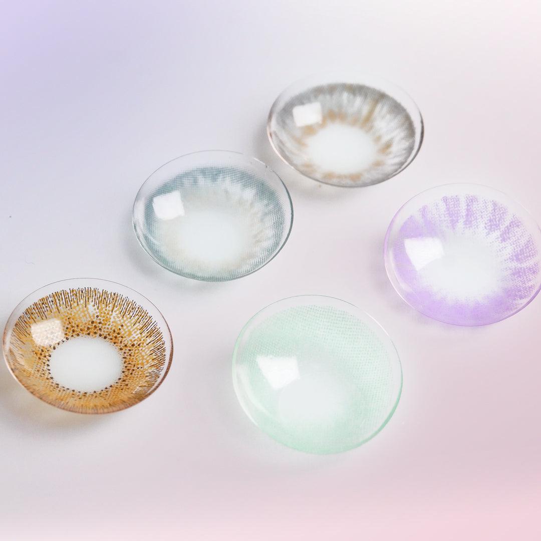 Various color contact lenses in blue, grey, violet, brown and pink, photographed on a purple background